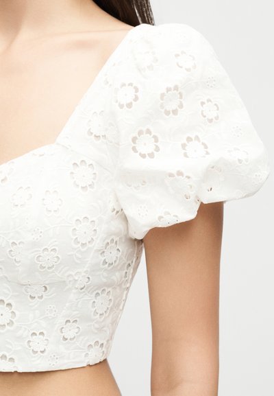 Crop top en coton blanc avec manches courtes bouffantes, broderie florale ajourée et encolure carrée sur une personne.