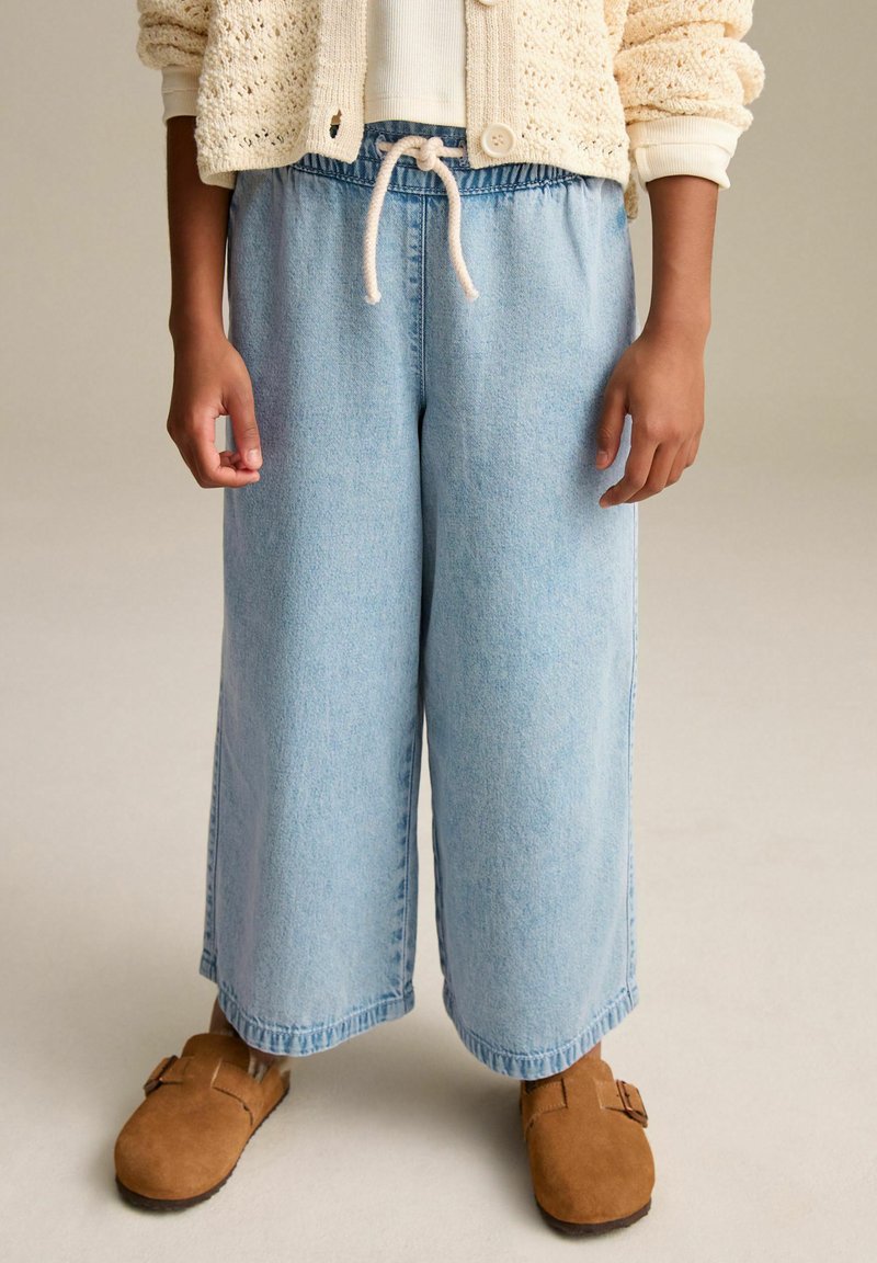 Lyseblå vidde jeans med elastisk talje og snoretræk, sammen med brune ruskindstræsko, sat mod en neutral baggrund.