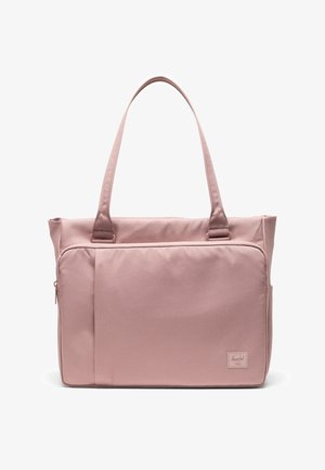 Sac cabas rose doux avec de longues poignées, poche zippée à l'avant et petite étiquette de marque rectangulaire en bas à droite.