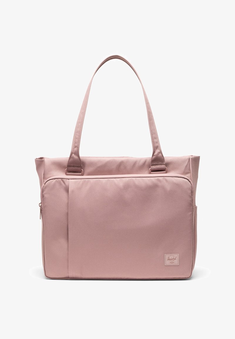 Mekana ružičasta torba za nošenje s dugim ručkama, prednjim džepom na zatvarač i malom pravokutnom oznakom brenda u donjem desnom kutu.
