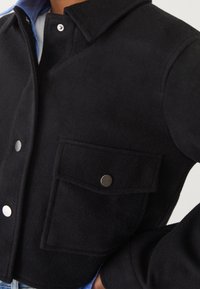 Černý vlněný kabát s velkou přední kapsou, stříbrnými patentkami, límcem, nošený přes modrou košili a světlé džínové kalhoty.