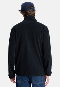 Giacca in pile nera con colletto alto, maniche lunghe e design semplice. Il tessuto ha una consistenza morbida, abbinata a jeans in denim blu.