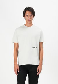 Lys grå bomulds t-shirt med rund hals, korte ærmer og et sort tekstlogo på venstre bryst. Glat tekstur, afslappet pasform.