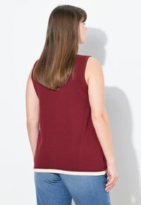 Vrouw met lang bruin haar, gekleed in een mouwloze donkerrode top met witte biezen en blauwe jeans, staand met haar gezicht naar een witte muur.