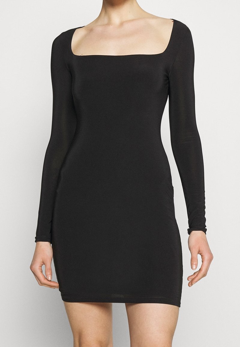 Femme portant une robe noire ajustée à manches longues avec un décolleté carré, debout devant un fond clair uni.