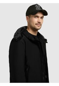 Black puffer jacket with a fur-lined collar, side pockets, and a front zipper. Paired with a black cap featuring a raised logo.