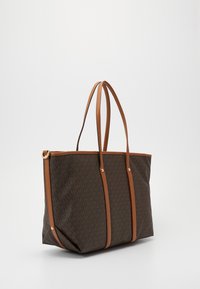 Sac fourre-tout marron foncé avec un motif de logo répétitif sur toute la surface, garniture en cuir beige, et deux longues poignées, sur un fond clair uni.