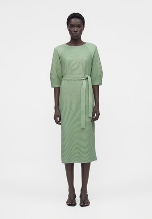 Kobieta w jasnooliwkowej, teksturowanej sukience midi z 3/4 rękawami i dopasowanym paskiem, nosząca czarne sandały na obcasie z paseczkami, stojąca na białym tle.