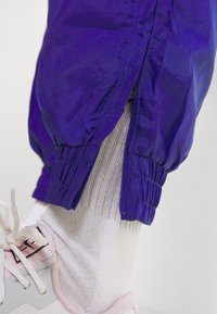 Fialová větrovka s elastickými manžetami a zipem u kotníku, doplněná bílou ponožkou a teniskou s růžovými detaily. Hladká textura.