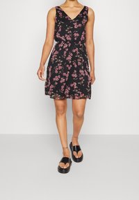 Schwarzes Blumenkleid mit V-Ausschnitt, ärmellosem Design und zarten rosa Blumenmustern. Getragen mit schwarzen Plateausandalen.
