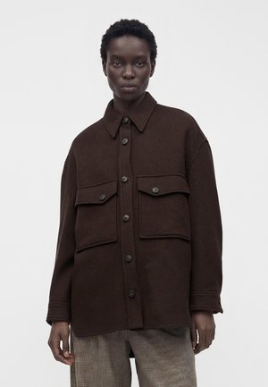 Μοντέλο φοράει ένα σκούρο καφέ oversized σακάκι με κουμπιά και μεγάλες τσέπες με καπάκια και καρό παντελόνια, στέκεται μπροστά σε ένα απλό φόντο.