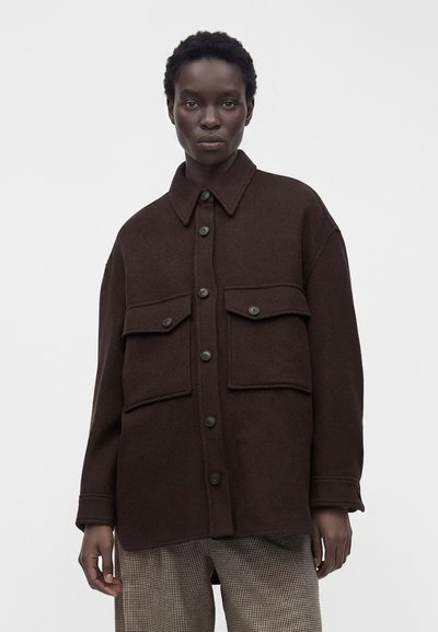 Mannequin portant une veste boutonnée oversize marron foncé avec de grandes poches à rabat et un pantalon à carreaux, debout devant un fond uni.