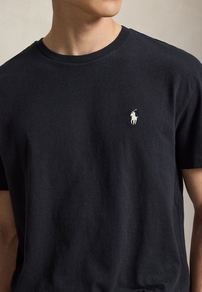 Uomo che indossa una t-shirt nera semplice con un piccolo logo bianco di un giocatore di polo sul petto sinistro, su uno sfondo neutro.