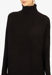 Pull noir à col roulé côtelé avec une coupe ample et une surface texturée. Présente un col montant et des manches larges pour une silhouette décontractée.