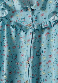 Blusa in tessuto azzurro chiaro con motivo floreale in accenti rosa, giallo e verde. Chiusura con bottoni e dettaglio colletto arricciato.
