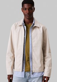 Giacca esterna beige chiaro con una texture liscia e chiusura con zip, indossata sopra una camicia verde e una camicia a righe blu.
