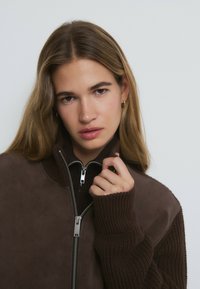 Veste zippée marron avec des manches côtel ées et une texture en daim lisse. Présente des éléments en métal argenté et un col haut.
