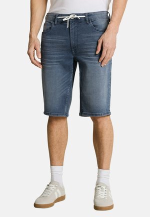 Hombre con pantalones cortos vaqueros azules con cordón blanco, calcetines blancos y zapatillas beige, de pie contra un fondo liso.