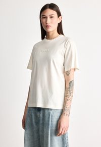 Μπεζ βαμβακερό t-shirt με κοντά μανίκια, με ανάγλυφο λογότυπο "BOSS" στο στήθος. Συνδυάζεται με ανοιχτό μπλε, φαρδιά τζιν παντελόνια.