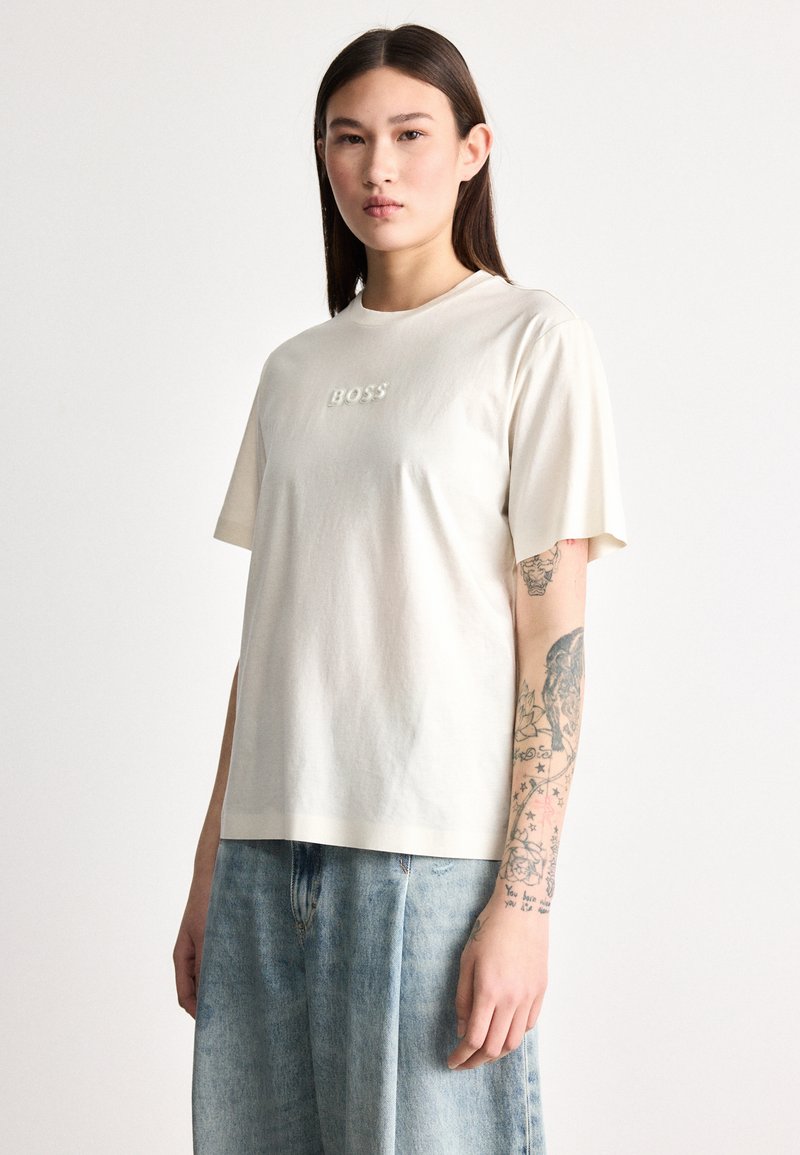 Μπεζ βαμβακερό t-shirt με κοντά μανίκια, με ανάγλυφο λογότυπο "BOSS" στο στήθος. Συνδυάζεται με ανοιχτό μπλε, φαρδιά τζιν παντελόνια.