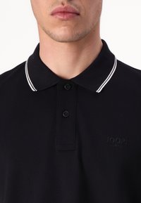 Čierne polo tričko s dvojitým gombíkovým zapínaním, bielo pruhovaným golierom a embosovaným logom na hrudi. Vyrobené z textúrovanej bavlny.