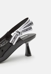 Scarpa slingback in pelle verniciata nera con tacco curvo, caratterizzata da un fiocco in tessuto bianco stampato con la scritta "KARL LAGERFELD" in nero.