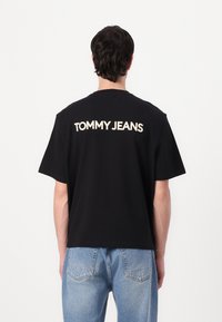 Svart bomull T-skjorte med "TOMMY JEANS" trykket i fet kremfarge på baksiden. Korte ermer og en avslappet passform, brukt med lyse blå jeans.