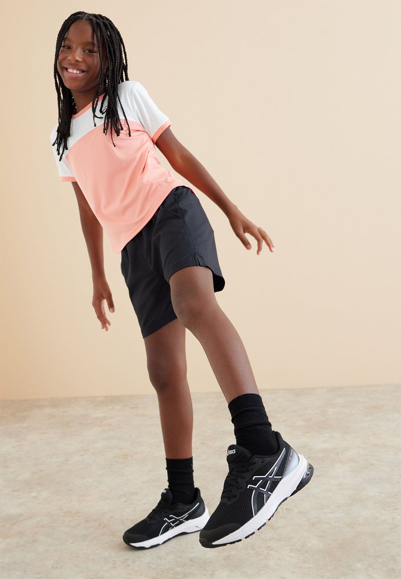 Camiseta atlética de manga corta en rosa y blanco combinada con pantalones cortos negros. Zapatillas negras y blancas con textura de malla y suelas acolchadas.