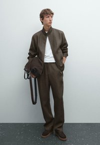 Brown textured jacket with a high collar, white shirt, brown trousers, and dark shoes. Holds a round brown bag with a strap.