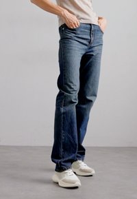 Denim jeans in dark blue, straight-leg design with visible stitching, front pockets, worn with light sneakers on a neutral background.