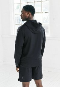 Musta urheiluhuppari, jossa on huppu, pitkät hihat ja rento istuvuus, yhdistetty mustiin shortseihin, joissa on heijastavia yksityiskohtia. Minimalistinen muotoilu.