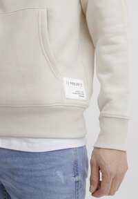 Bežová mikina s predným vreckom a rebrovanými manžetami, s oznámením "11 PROJECT" a detailami. Pod ňou je biele tričko.
