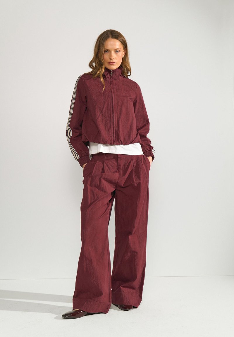 Femme portant un survêtement bordeaux avec des bandes blanches sur les manches, pantalon à jambes larges, chemise blanche en dessous, debout les mains dans les poches.