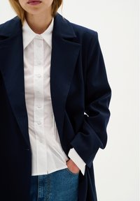 Cappotto blu navy con un design sartoriale, caratterizzato da un colletto a punta, indossato sopra una camicia bianca con bottoni. Jeans in denim visibili in vita.