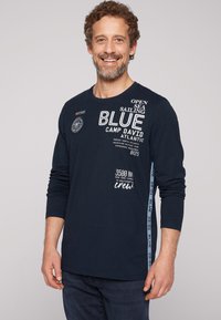 Maglietta navy a maniche lunghe con testo bianco, un'etichetta circolare e dettagli laterali. Realizzata in un tessuto liscio con un design a vestibilità regolare.