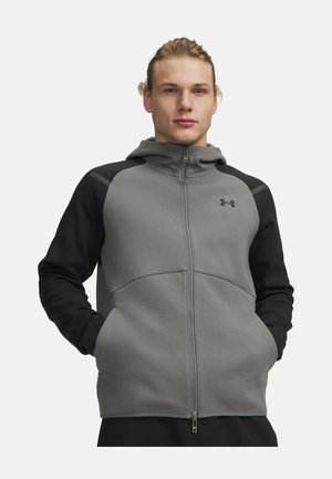 Man draagt een grijs-zwarte zip-up hoodie met capuchon, staat met handen in de zakken tegen een effen achtergrond.