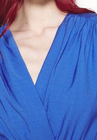 Personne aux cheveux roux portant un haut bleu texturé à col en V, montrant l'encolure et le haut de la poitrine.