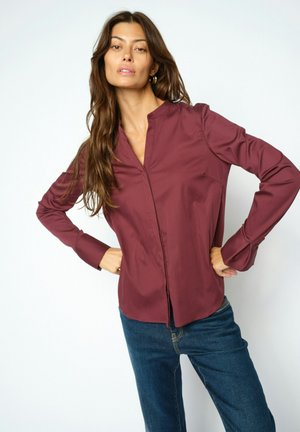 Vrouw met lang bruin haar, gekleed in een bordeauxrode longsleeve en blauwe spijkerbroek, staand met handen in de zij tegen een witte achtergrond.