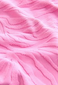 Mīksta rozā auduma ar reljefainiem ģeometriskiem rakstiem un redzamām pīšanas detaļām, radot taustāmu virsmu.