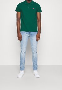 Homme portant un polo vert foncé, un jean bleu clair et des baskets blanches, debout devant un fond blanc uni.