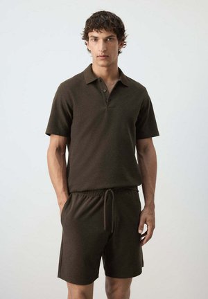 Hombre de pie vestido con una camiseta polo de manga corta marrón oscuro y pantalones cortos a juego con cordón, frente a un fondo claro y sencillo.