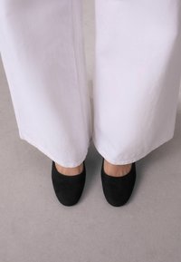 Zapatos negros de ante de puntera cerrada con una textura suave, combinados con pantalones anchos blancos que cubren parcialmente las aberturas de los zapatos, revelando piel desnuda en los tobillos.