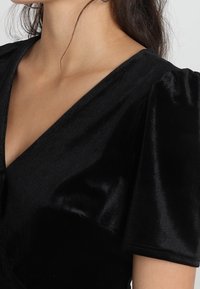 Czarny aksamitny top z głębokim dekoltem w kształcie litery V, krótkimi bufkami na rękawach oraz miękką, gładką fakturą. Materiał wygląda na błyszczący i luksusowy.