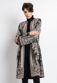 Lang sort cardigan med indviklede hvide og røde mønstre, med høj krave og brede ærmer, lavet af struktureret stof.