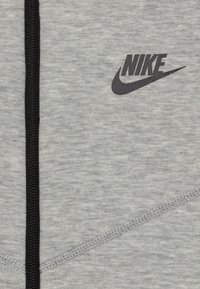 Šedá mikina s texturovanou tkaninou, s černou lemovkou na zipu a výrazným černým logem Nike na přední straně.