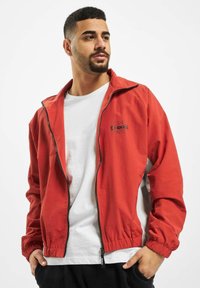 Jachetă roșie din material impermeabil, cu fermoar în față, manșete elastice și detalii cu logo. Puritată peste un tricou alb, asociată cu pantaloni negri.