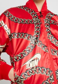 Röd satin bomberjacka med ett kedjemönster i svart och guld, med dragkedja framtill och ribbade ärmslut och krage. Nike-logotypen finns på jackan.