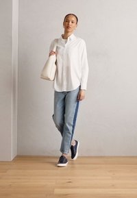 Vit skjorta med knäppning, ljusblå jeans med mörka sidostråk, marinblå sneakers och en krämfärgad axelväska på en träbotten mot en grå vägg.