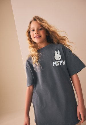 Ung pige med langt, bølget blondt hår iført en løs mørkegrå T-shirt med en hvid tegneseriekanin og "MIFFY" tekst.