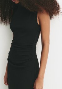 Frau mit lockigem Haar, die ein ärmelloses, tailliertes schwarzes Kleid trägt und vor einer weißen Wand steht, von Hals bis Oberschenkel gezeigt.